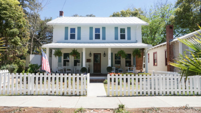 Exterior view of a finished home with a new roof and white siding with blue window storm shutters with a small gate and walkway to the home.