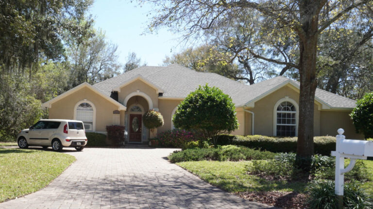 Exterior view of a finished home with a new roof surrounded by grass, drive-way and trees.