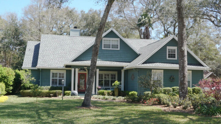 Exterior view of a finished home with new roofing and siding surrounded by grass, trees and bushes.