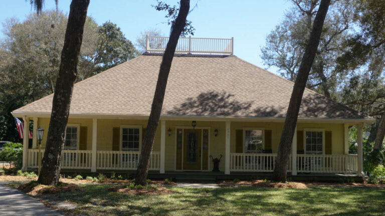 Exterior view of a finished home with a new roof and viewing deck on top of the rood. The home is also surrounded by three large trees in front with a grass lawn.